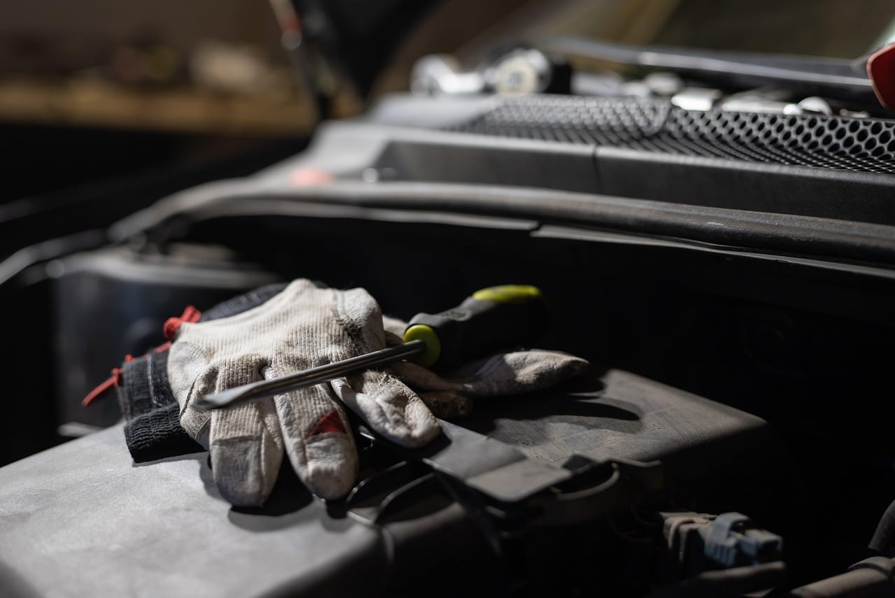 Un paio di guanti da lavoro sporchi e un cacciavite appoggiati sul motore di un'auto con il cofano aperto, in penombra.