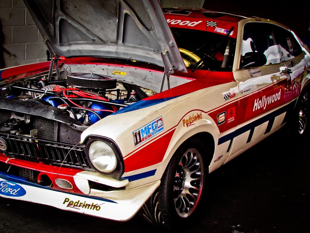 Auto da corsa d'epoca bianca, rossa e blu con scritte Ford e Hollywood, cofano aperto che mostra il motore, in un garage.