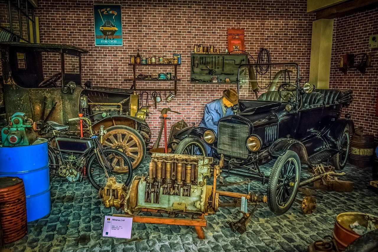 Officina d'epoca con muro di mattoni: auto nera scoperta, un manichino in tuta blu china sul motore, moto antica e barili.