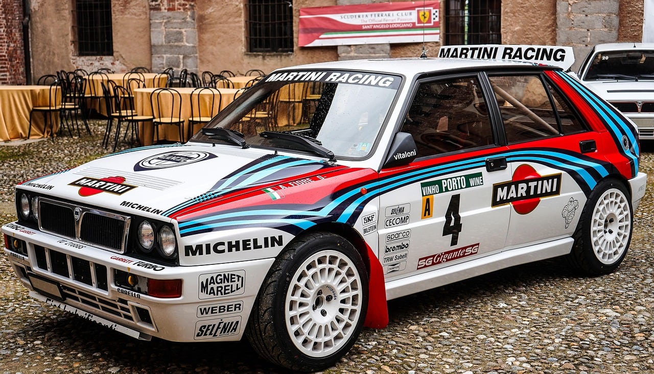 Auto da rally bianca con livrea Martini Racing a strisce rosse e blu, numero 4, parcheggiata su un selciato davanti a tavoli di un ristorante.