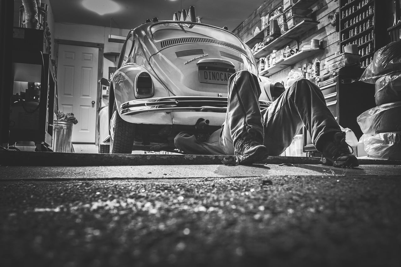 In un garage, un meccanico sdraiato sotto un Maggiolino d'epoca; si vedono solo gambe e scarponi. Foto in bianco e nero.