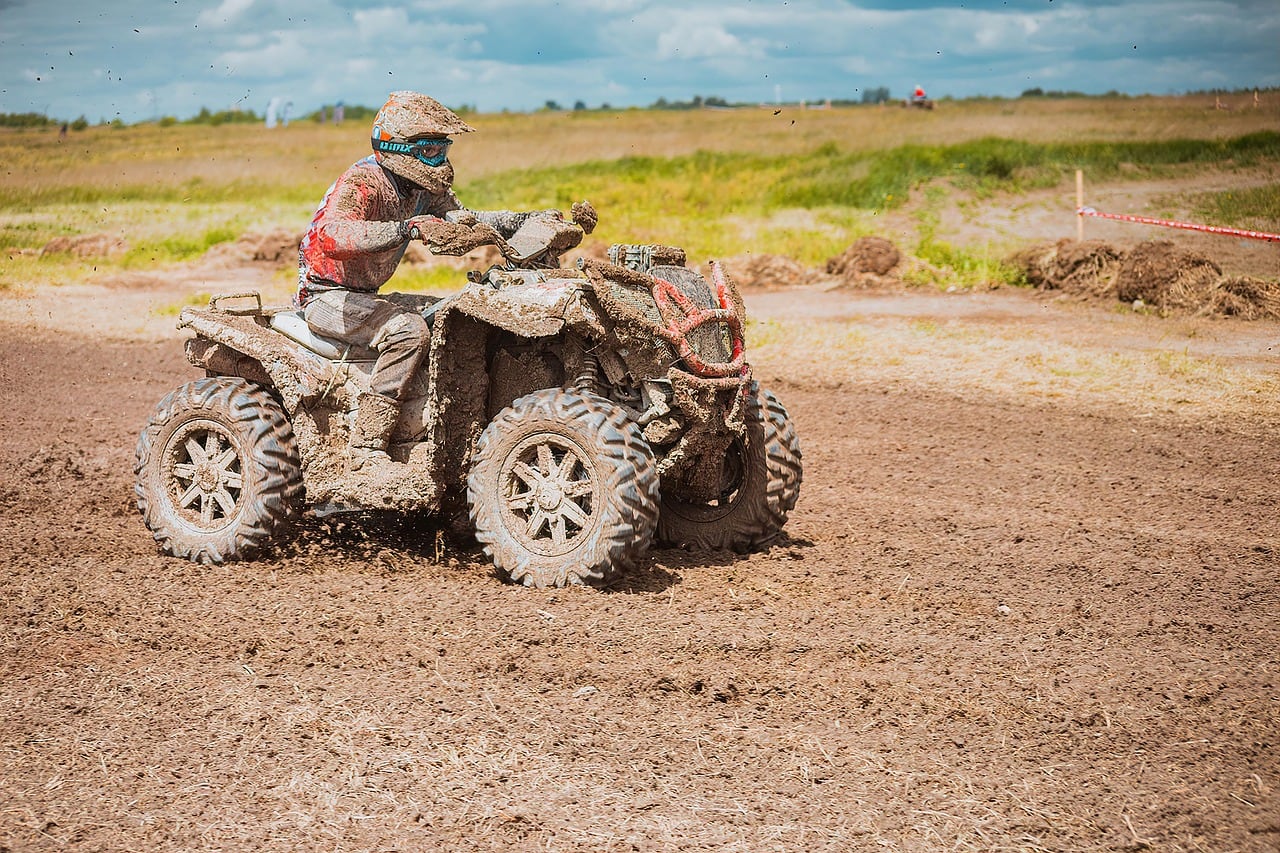 Pilota con casco su un quad coperto di fango che percorre un tracciato di terra battuta in mezzo ai campi.