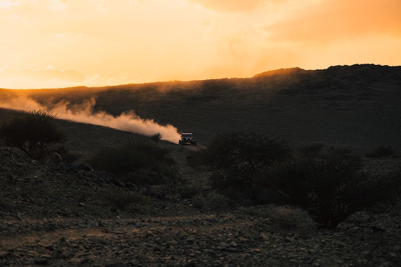 Un fuoristrada percorre una pista sterrata in un paesaggio roccioso al tramonto, sollevando una lunga scia di polvere sotto un cielo arancione.